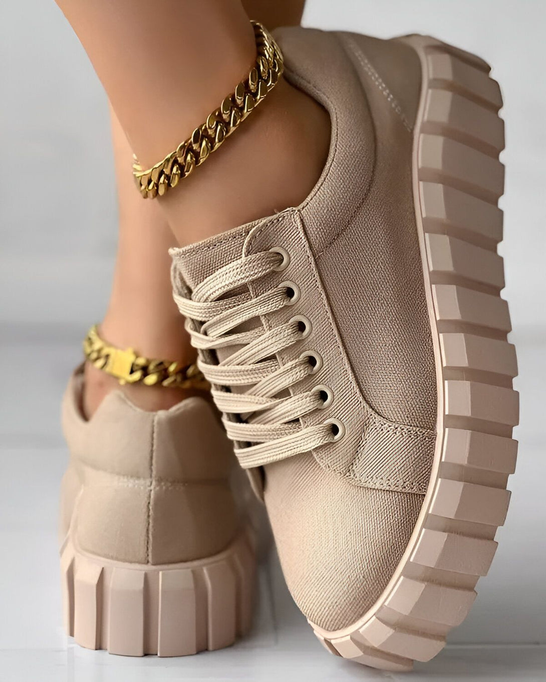 Une image d'une paire de chaussures beiges avec des lacets beiges et une semelle beige. Il y a une chaîne de cheville en or sur la cheville droite du modèle.