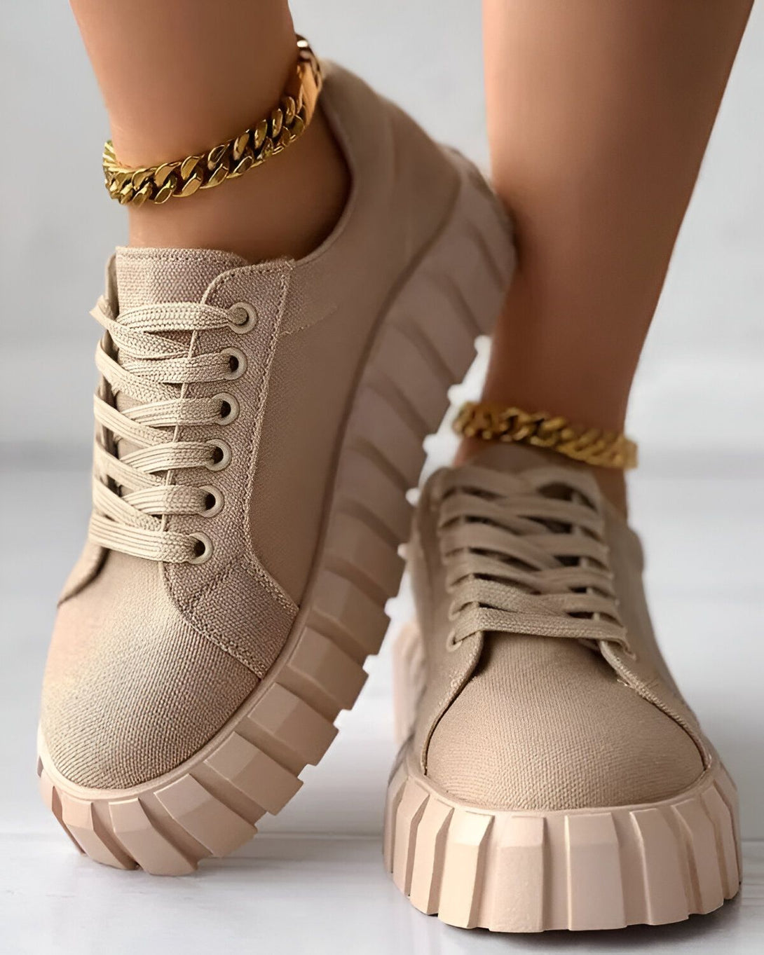 Une paire de baskets beiges avec des lacets et une semelle épaisse. Les baskets sont portées par une femme qui porte également un bracelet de cheville en or.