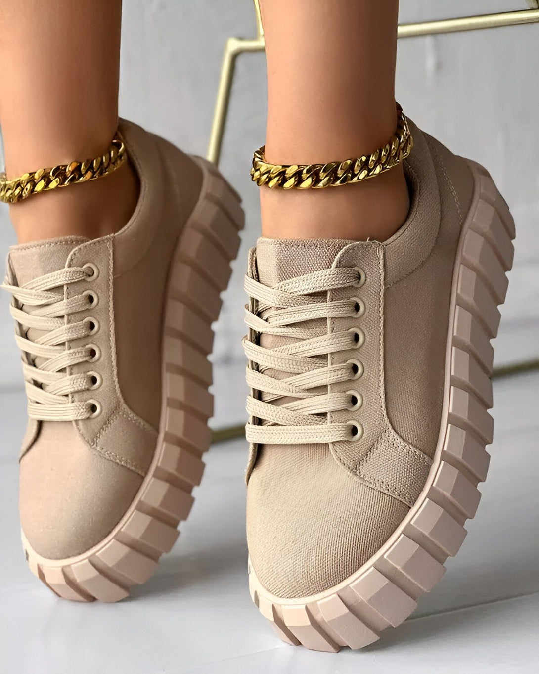 Une paire de baskets beiges avec des lacets beiges et une semelle épaisse beige et rose. Les baskets sont portées par une femme qui porte également une chaîne de cheville en or.