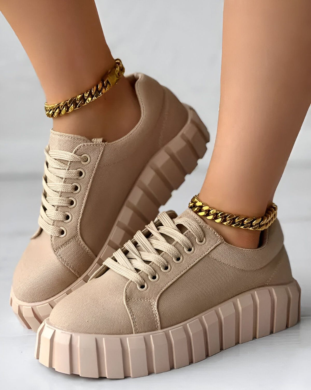 Une paire de baskets beiges avec des lacets beiges et une semelle épaisse beige. Les baskets sont portées par une femme qui a une chaîne en or autour de la cheville.