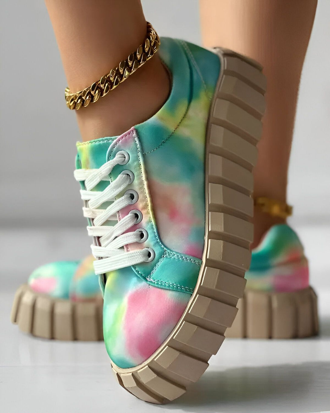 Une image d'une paire de chaussures de sport multicolores avec des lacets blancs et une semelle épaisse. Les chaussures sont portées par une femme qui porte également une chaîne de cheville en or.