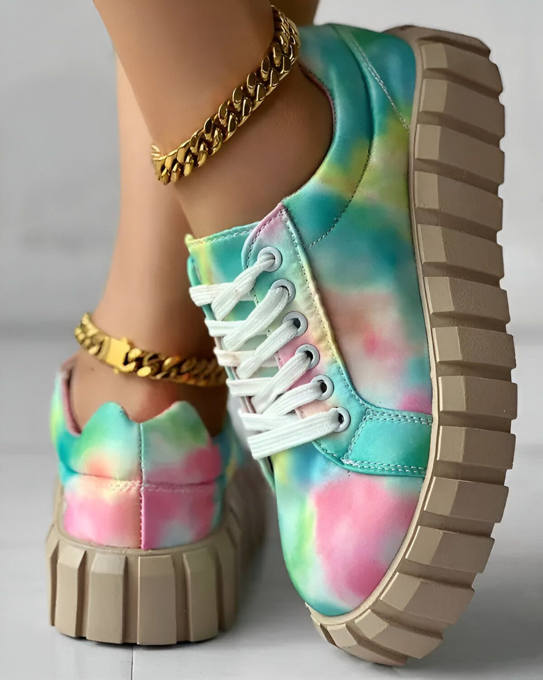 Une femme porte des baskets tie-dye roses, bleues et vertes avec des lacets blancs et une semelle épaisse beige. Elle porte également une chaîne de cheville en or.
