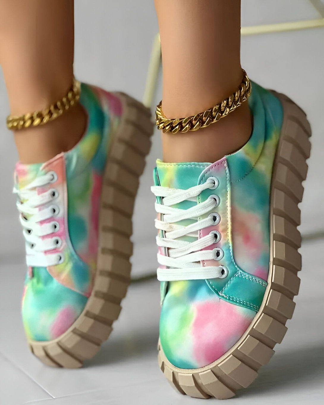 Une femme porte des baskets tie-dye colorées avec des lacets blancs et une semelle épaisse. Elle porte également une chaîne de cheville en or.