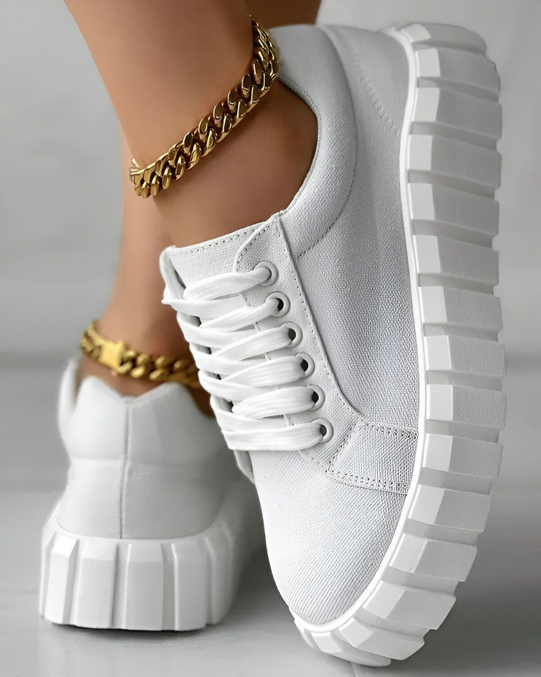 Une image d'une paire de baskets blanches avec une semelle épaisse. Les baskets sont lacées et ont une languette blanche. La personne qui porte les baskets a une cheville fine et porte un bracelet de cheville en or.