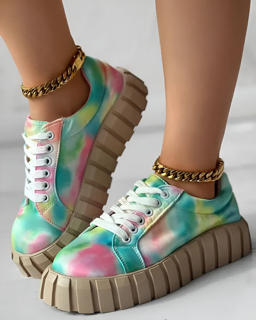 Une femme porte des baskets tie-dye colorées avec des lacets blancs et une semelle épaisse. Elle porte également un bracelet de cheville en chaîne dorée.