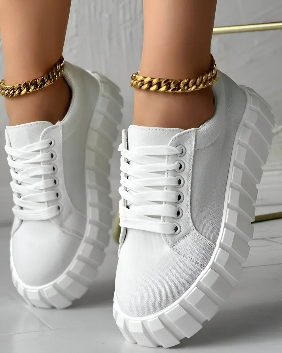 Une paire de baskets blanches à lacets avec une semelle épaisse. Les baskets sont portées par une femme qui porte également une chaîne en or autour de la cheville.