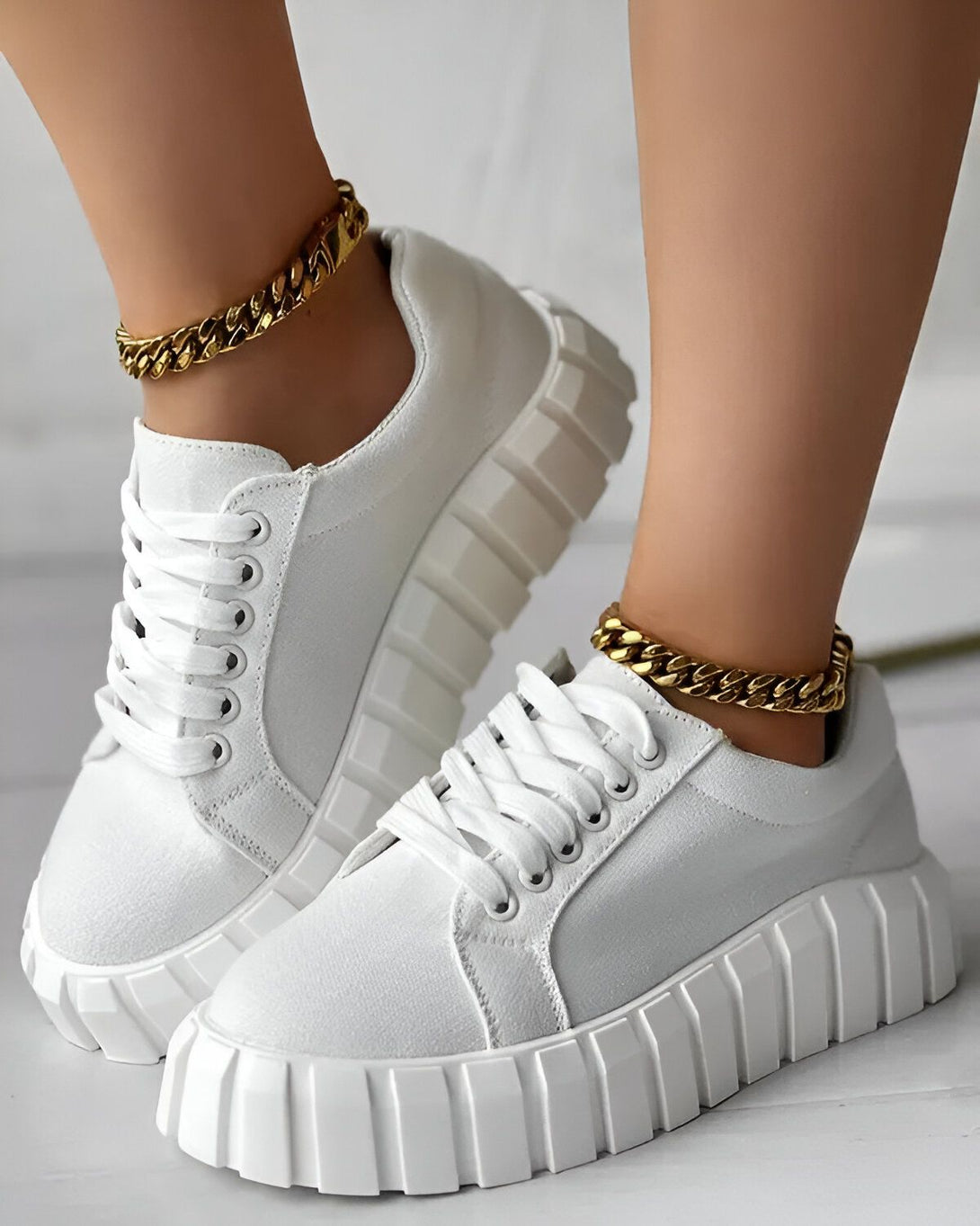 Une femme porte des baskets blanches avec des lacets blancs et une semelle épaisse. Elle porte également une chaîne en or autour de la cheville.