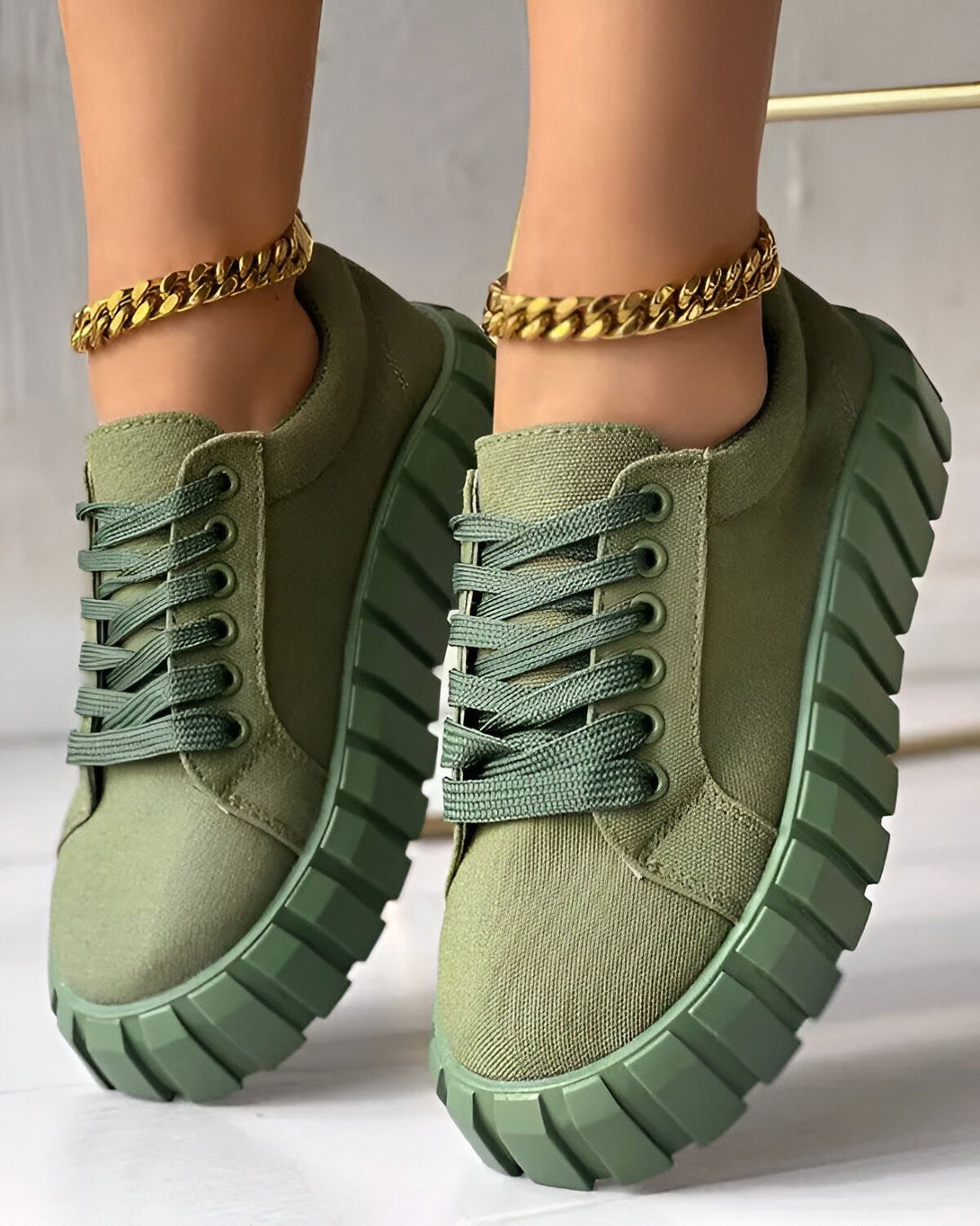 Une paire de baskets vertes avec des lacets verts et une semelle épaisse. Les baskets sont portées par une femme qui a une chaîne en or autour de la cheville.