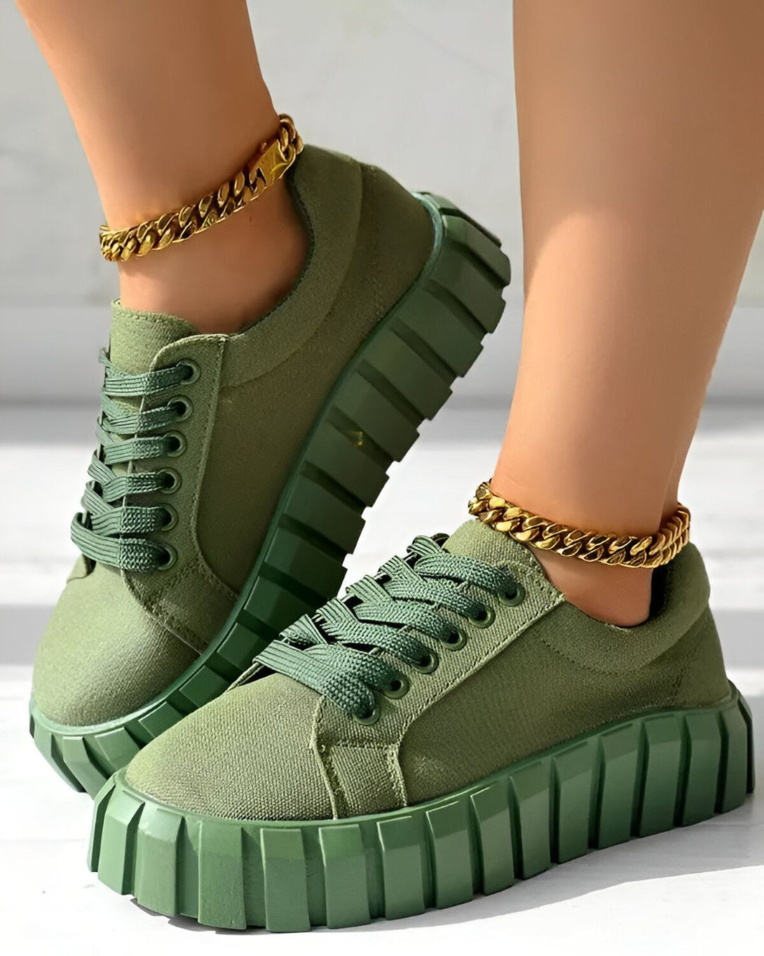 Une paire de baskets vertes avec des lacets et une semelle épaisse. Les baskets sont portées par une femme qui porte également des bracelets de cheville en or.