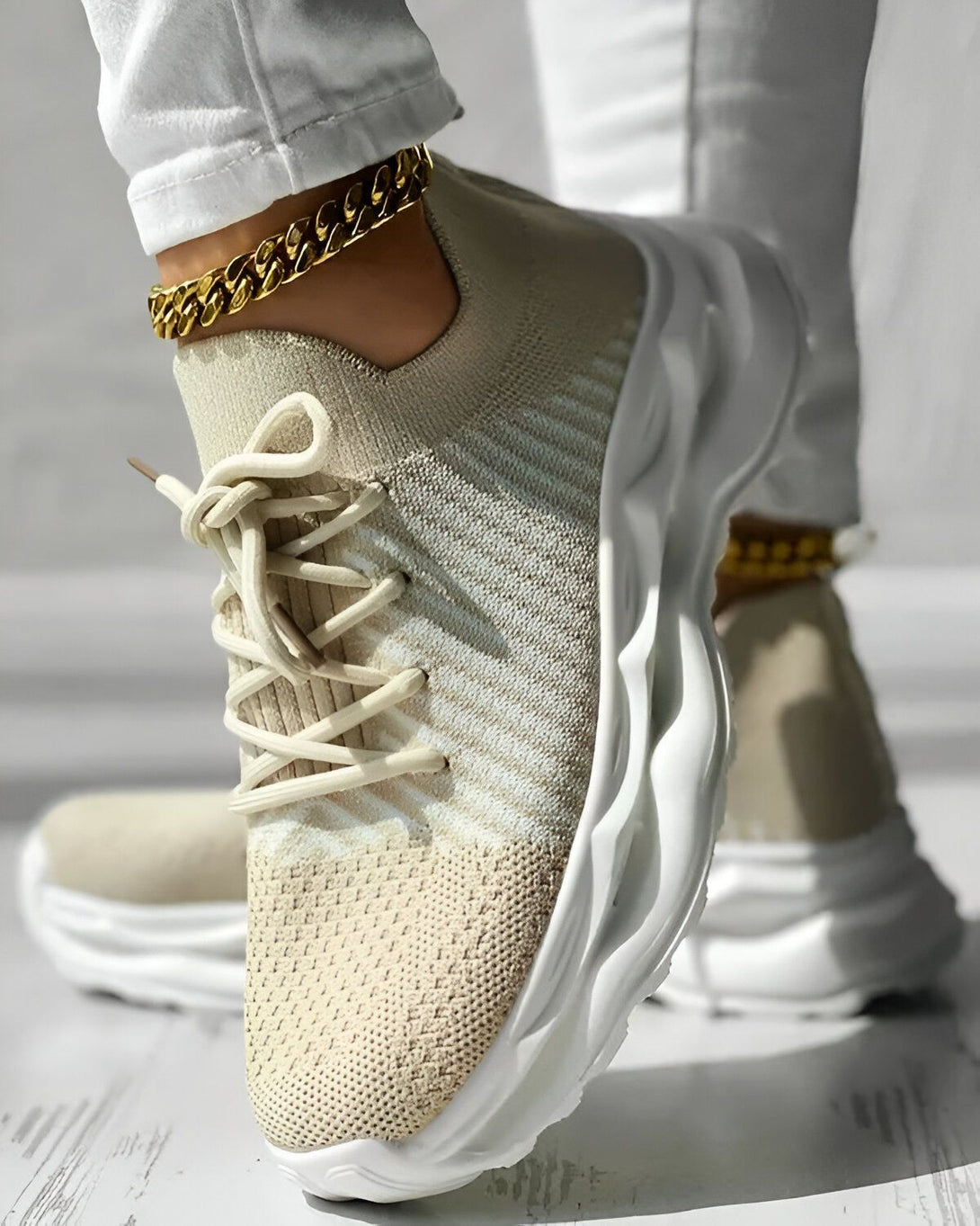 Une femme porte des baskets beiges et blanches avec des lacets beiges. Elle porte également un jean blanc et une cheville dorée. Les baskets sont en maille et ont une semelle épaisse.