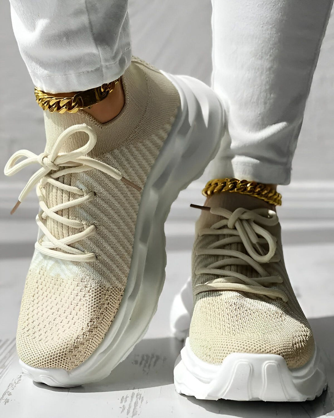 Une femme porte des baskets beiges avec des lacets blancs et des semelles blanches. Elle porte également un pantalon de jogging blanc et des chaussettes blanches. Les baskets sont de la marque Nike et ont une grosse semelle. La femme est debout sur un sol en béton.