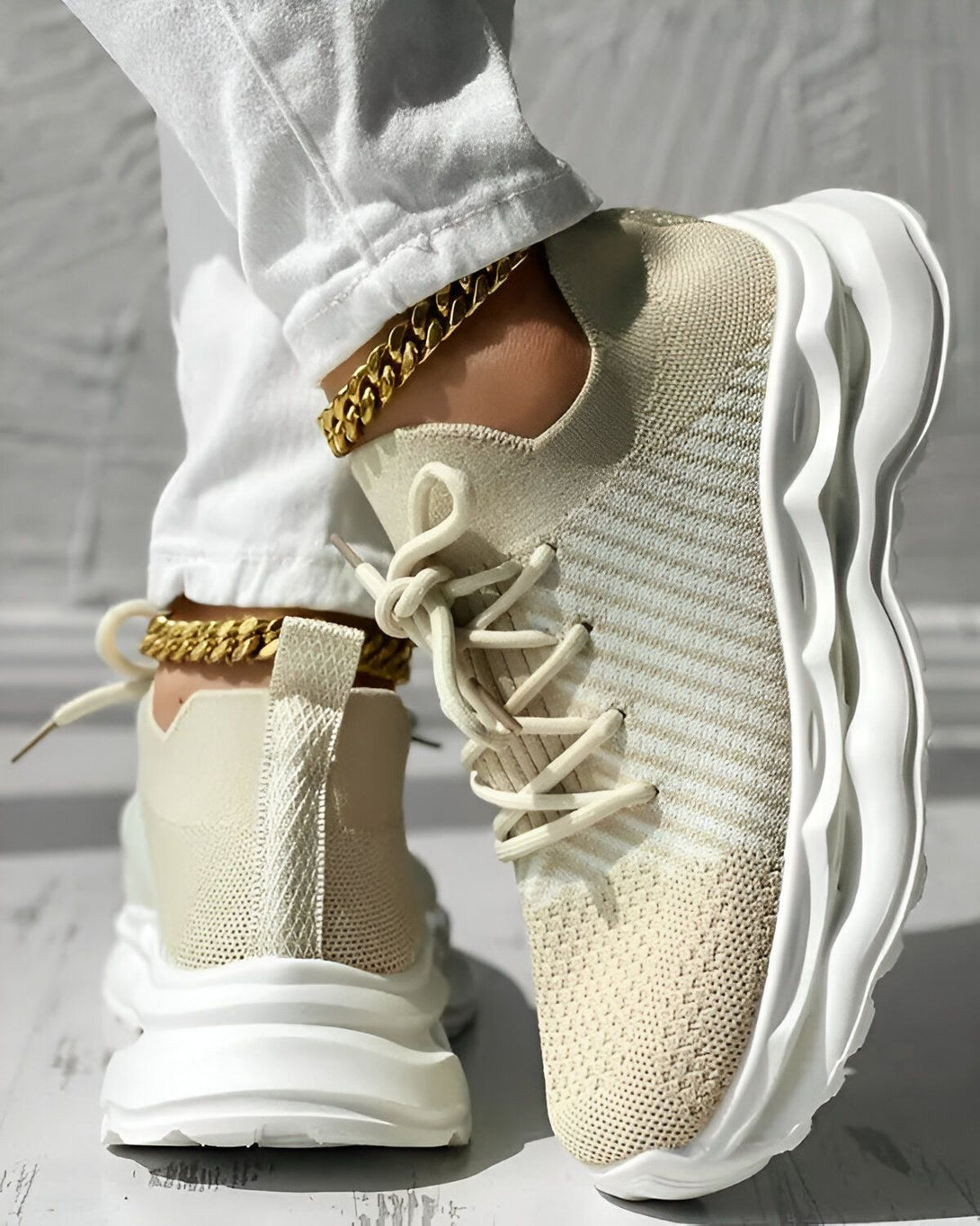 Une femme porte des baskets beiges et blanches avec des lacets beiges. Elle porte également un jean blanc et un bracelet de cheville doré.