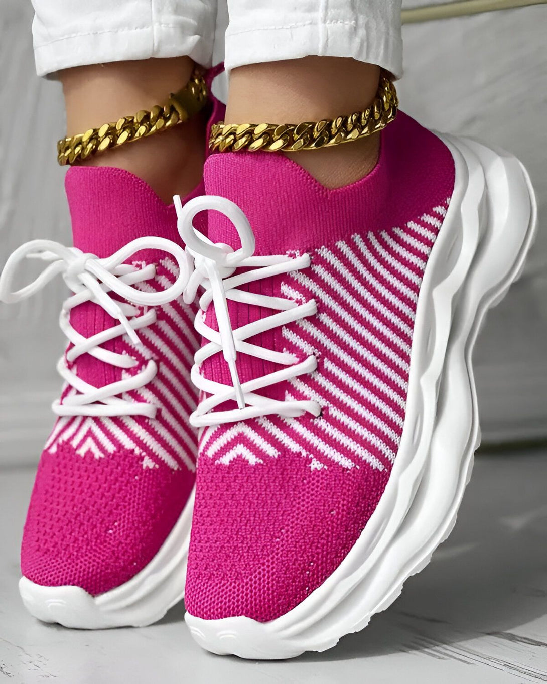Une femme porte des baskets roses et blanches. Les baskets sont en maille et ont des lacets blancs. La femme porte également un pantalon blanc et des chaussettes blanches. Elle est debout sur un sol en bois.