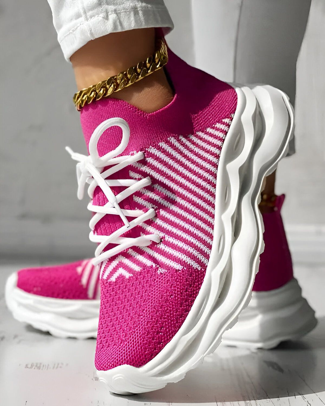 Une femme porte des baskets roses et blanches. Les baskets sont en maille et ont des lacets blancs. La semelle est blanche et épaisse. La femme porte également un pantalon blanc et un bracelet doré à la cheville.