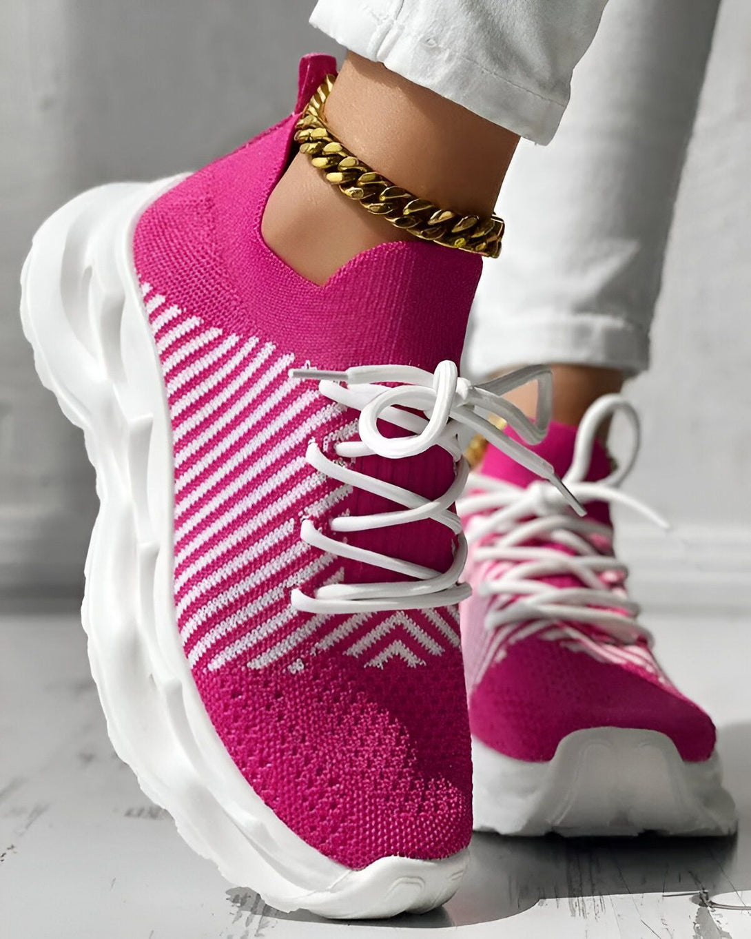 Une femme porte des baskets roses et blanches. Les baskets sont en maille et ont des lacets blancs. La femme porte également un jean blanc et un bracelet doré à la cheville.