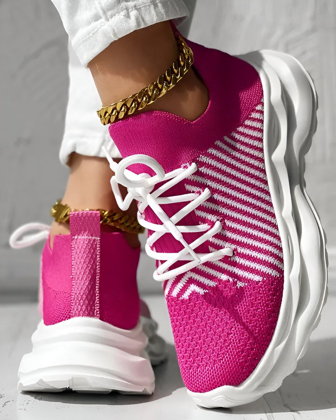 Une femme porte des baskets roses et blanches. Les baskets sont lacées avec des lacets blancs et ont une semelle blanche. La femme porte également un pantalon blanc et un bracelet de cheville doré.