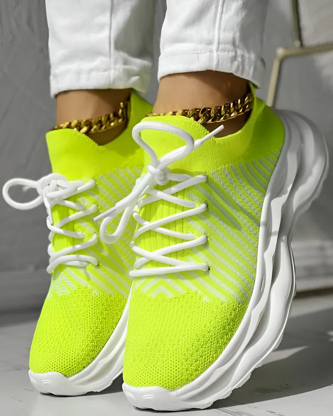 Une femme porte des baskets vert fluo avec des lacets blancs. Les baskets ont une semelle blanche et des rayures blanches. La femme porte également un pantalon blanc et un collier doré.