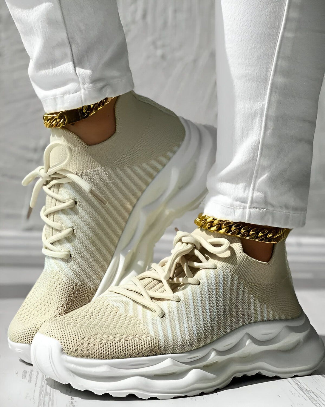 Une femme porte des baskets beiges et blanches avec des lacets beiges. Elle porte également un jean blanc et des bracelets de cheville dorés.