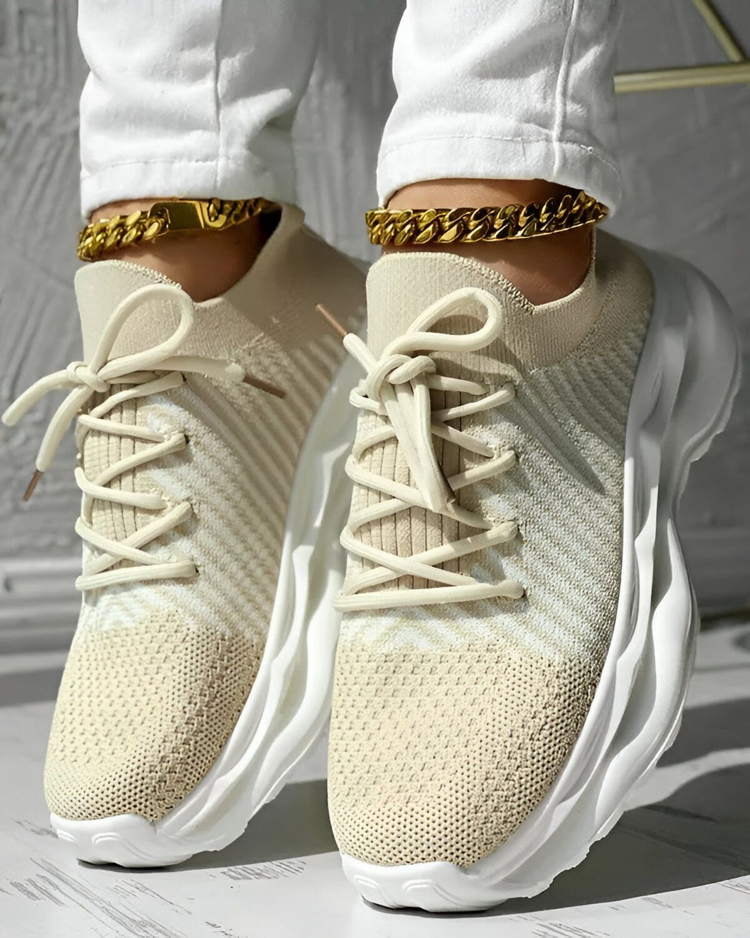 Une femme porte des baskets beiges et blanches avec des lacets beiges. Elle porte également un pantalon blanc et un bracelet de cheville doré.