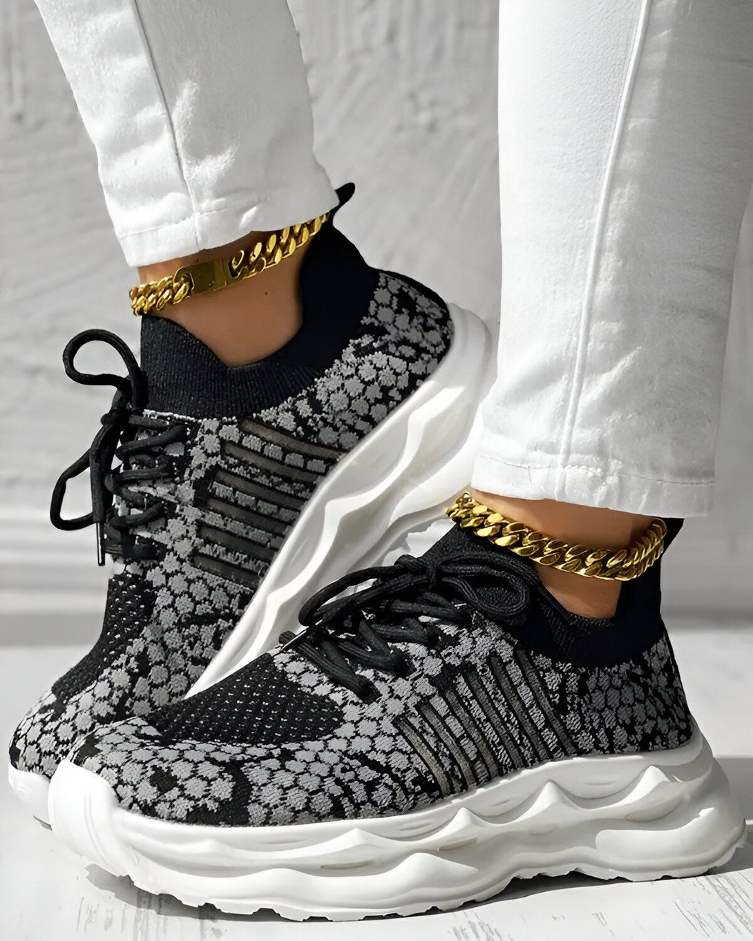 Une femme porte des baskets noires et grises avec des lacets noirs. Elle porte également un pantalon blanc et des chaussettes blanches. Les baskets ont une semelle blanche et une tige en tissu noir et gris. La femme est debout sur un sol en bois.