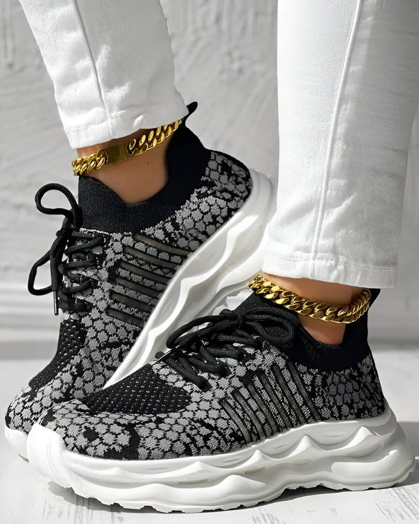 Une femme porte des baskets noires et grises avec des lacets noirs. Elle porte également un pantalon blanc et des chaussettes blanches. Les baskets ont une semelle blanche et une tige en tissu noir et gris. La femme est debout sur un sol en bois.