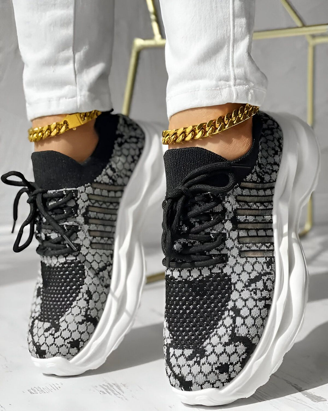 Une femme porte des baskets noires et grises avec des lacets noirs. Elle porte également un jean blanc et des bijoux en or.