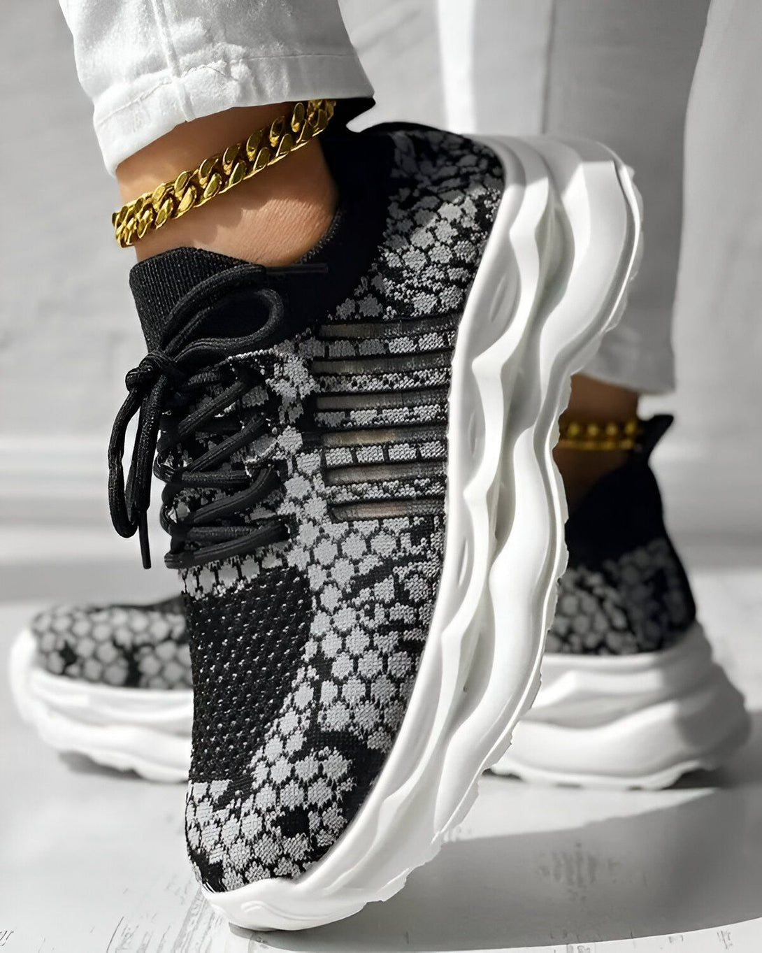 Une femme porte des baskets noires et grises avec une semelle blanche. Elle porte également un jean blanc et un bracelet de cheville doré.