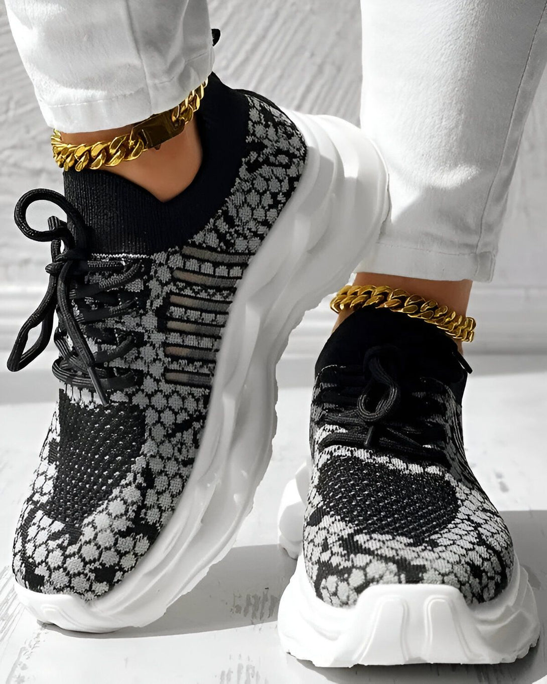 Une femme porte des baskets noires et grises avec des lacets noirs. Elle porte également un pantalon blanc et des chaussettes blanches. Les baskets ont une semelle blanche et une tige en mesh noir et gris. La femme est debout sur un sol en béton.