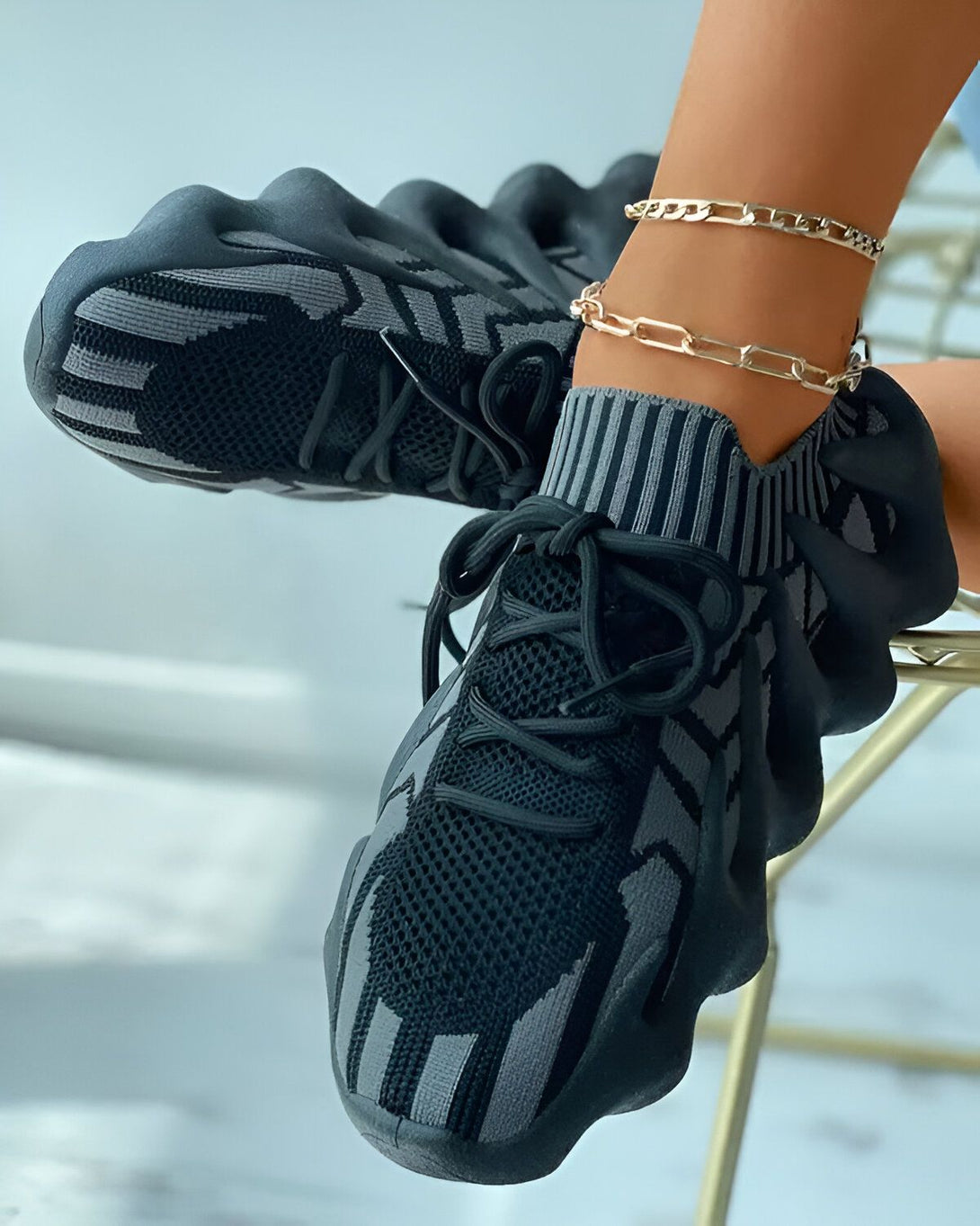 Une femme porte des baskets noires et grises. Les baskets sont lacées et ont une semelle épaisse. La femme est assise sur une chaise et porte une robe grise.