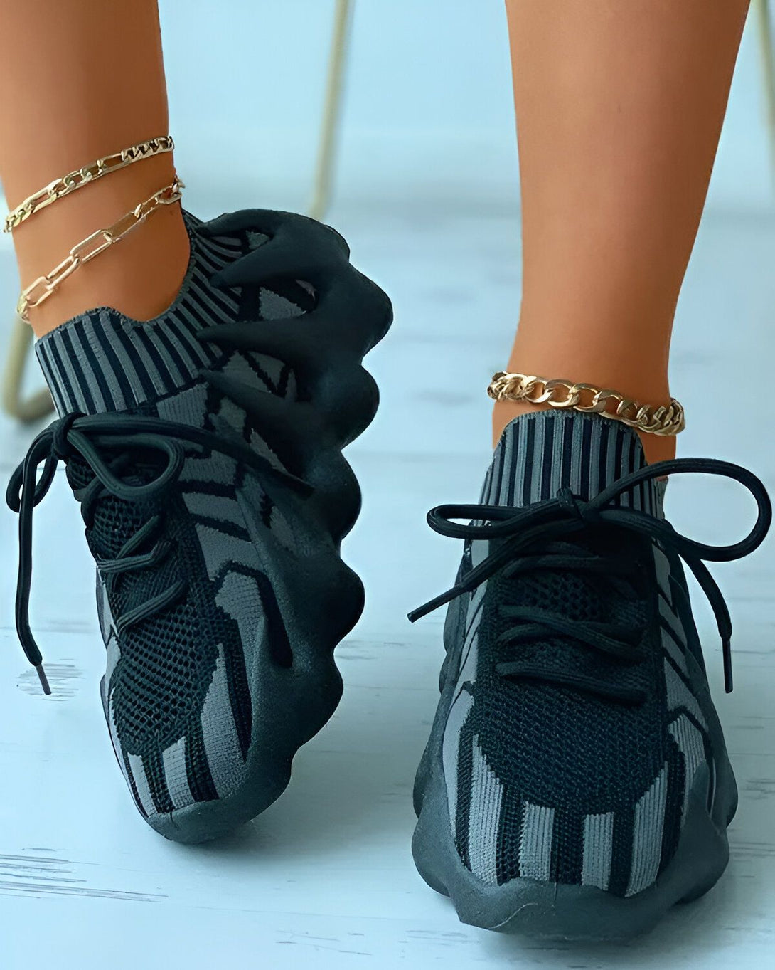 Une paire de baskets noires et grises à lacets avec une semelle épaisse. Les baskets sont portées par une femme qui porte également des bracelets de cheville en chaîne dorée.