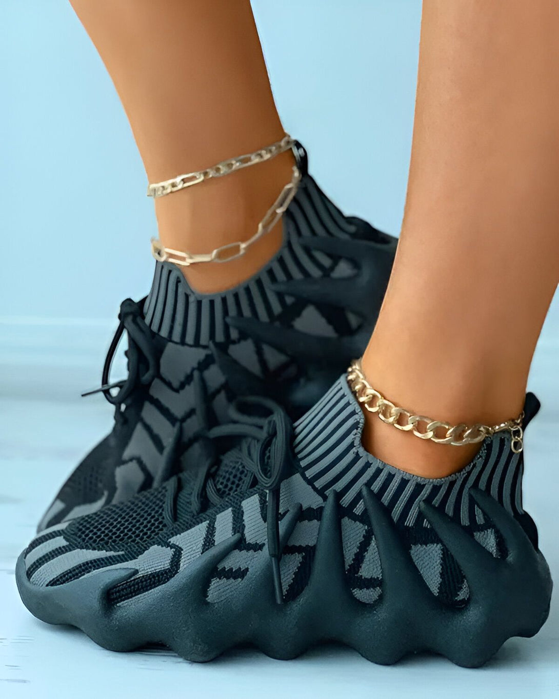 Une femme porte des baskets noires et grises avec des lacets et des semelles épaisses. Elle porte également des chaussettes noires et des bijoux aux pieds.
