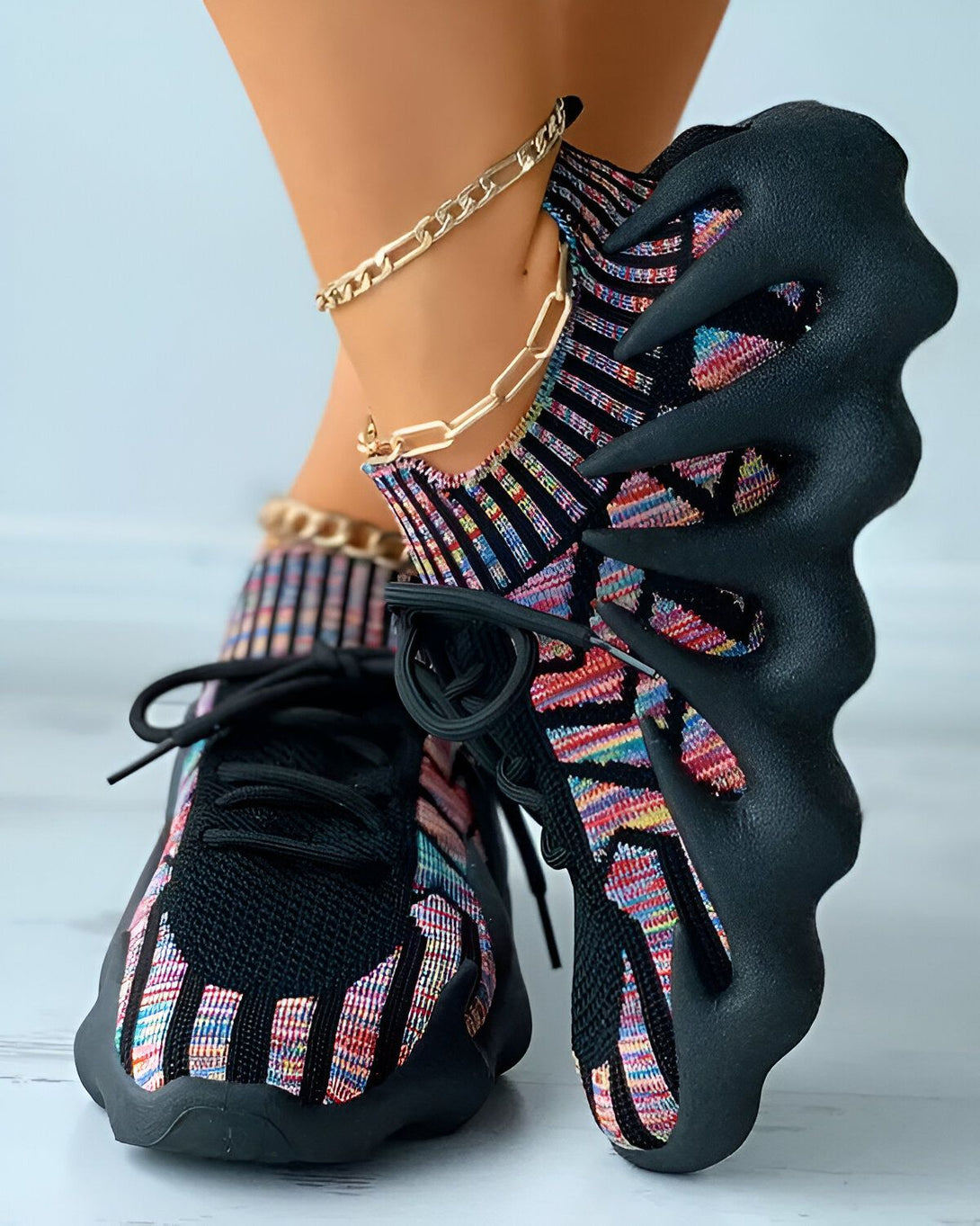 Une femme porte des baskets noires et multicolores. Les baskets sont lacées et ont une semelle épaisse. La femme porte également une chaîne dorée à la cheville.