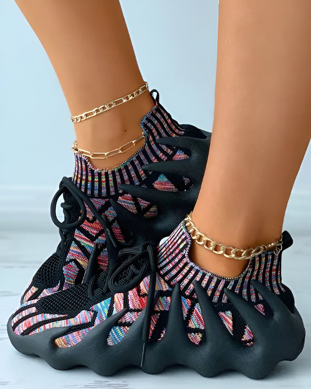Une femme porte des baskets noires et multicolores. Les baskets sont enfilées et les lacets sont noirs. La femme porte également deux chaînes dorées autour de la cheville.
