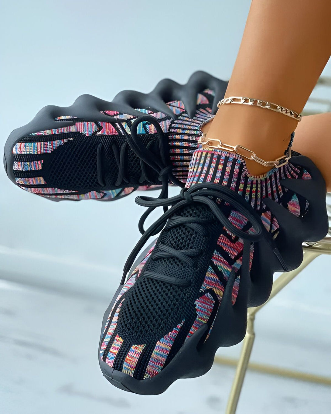 Une femme porte des baskets noires et multicolores. Les baskets sont en maille et ont des lacets. La femme est assise sur une chaise et porte un pantalon noir et un haut blanc. Elle a une chaîne dorée à la cheville.
