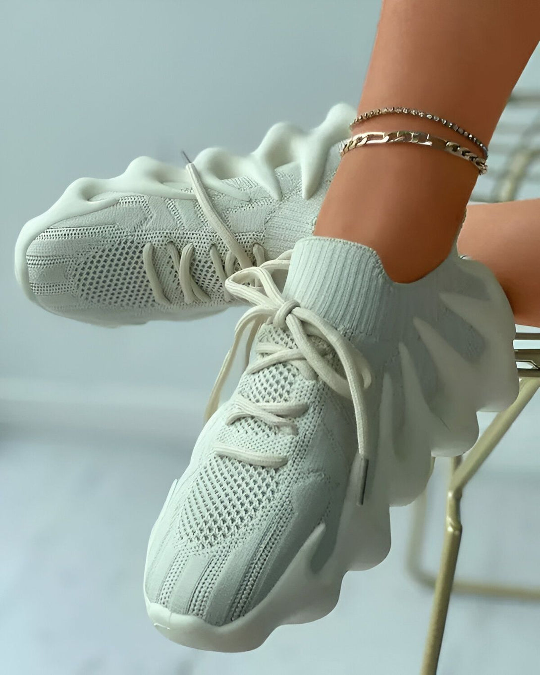 Une paire de baskets blanches avec des lacets blancs sur un pied et un bracelet de cheville en argent sur l'autre pied.
