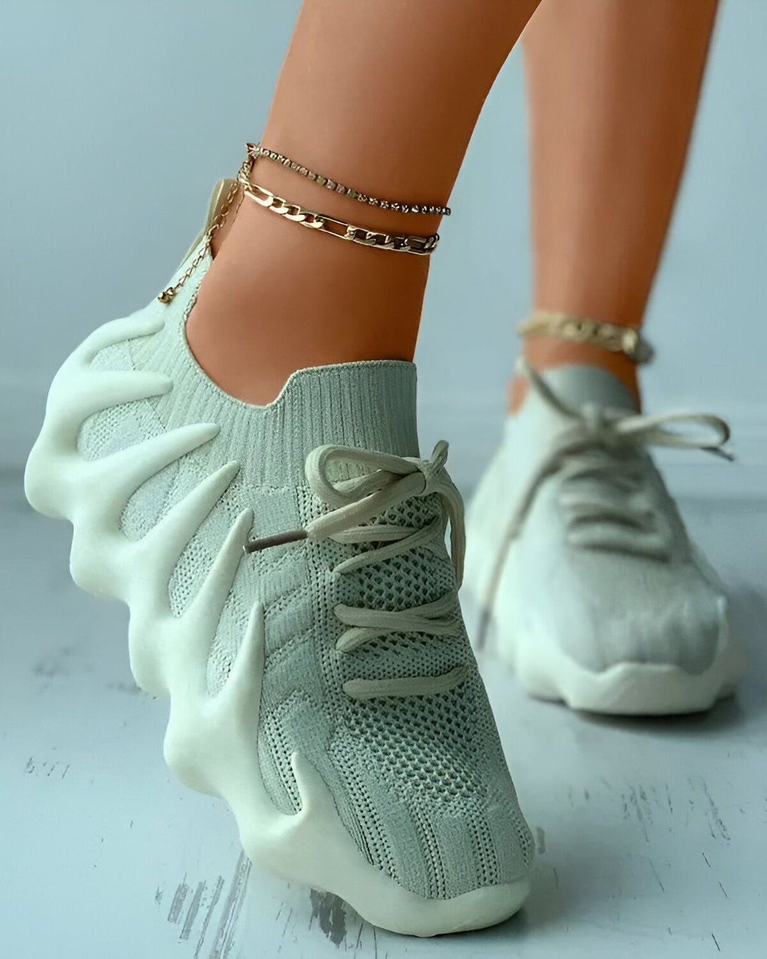 Une femme porte des baskets vert clair avec des lacets blancs. Les baskets sont en maille et ont une semelle épaisse. La femme porte également des bracelets de cheville en or.