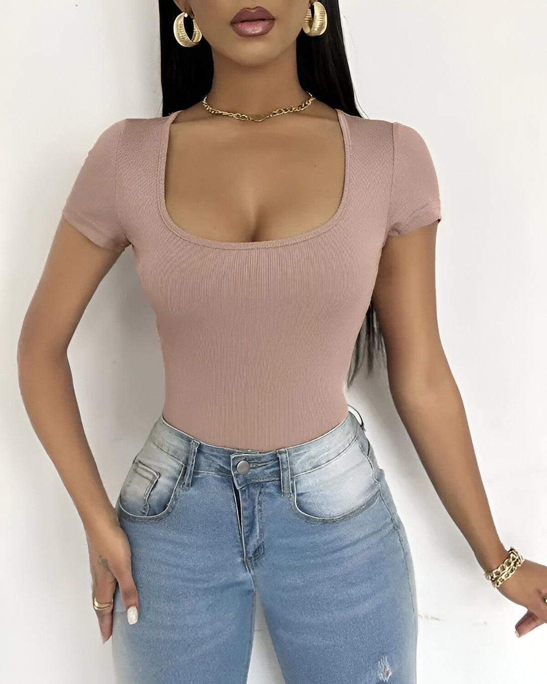 Une jeune femme porte un body moulant marron clair à manches courtes et un jean bleu taille haute. Elle a les cheveux longs noirs, un collier doré et des boucles d'oreilles dorées. Elle est maquillée et a les ongles manucurés.