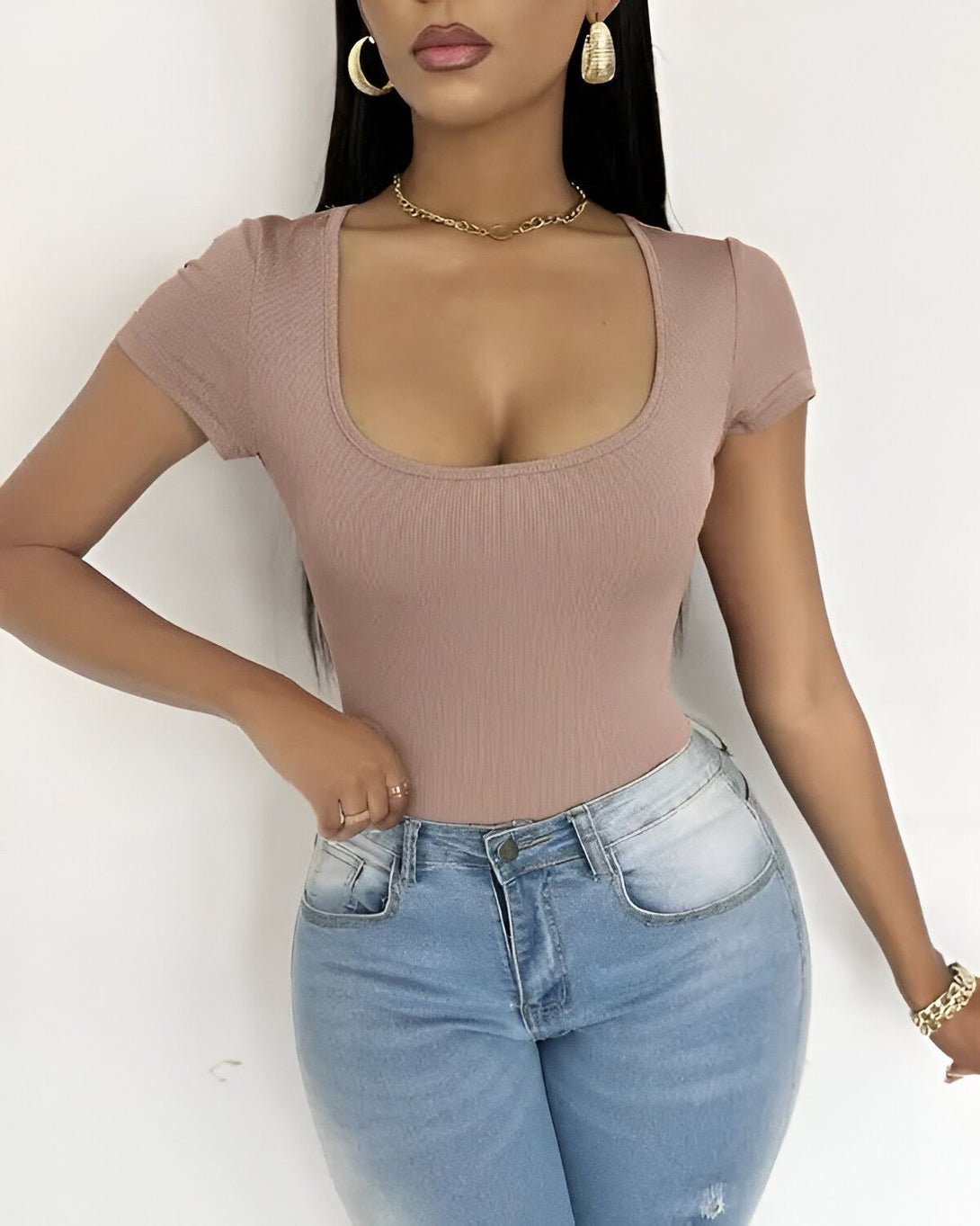 Une jeune femme porte un body moulant marron clair à manches courtes et un jean bleu troué taille haute. Elle a les cheveux longs noirs, un collier doré et des boucles d'oreilles dorées. Elle est maquillée et porte du gloss.