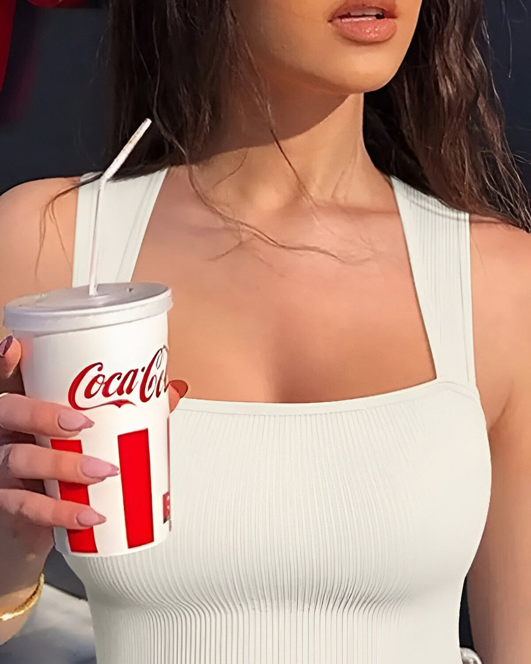 Une jeune femme tient une canette de Coca-Cola à la main. Elle porte un débardeur blanc et un collier doré. Ses ongles sont peints en rose.
