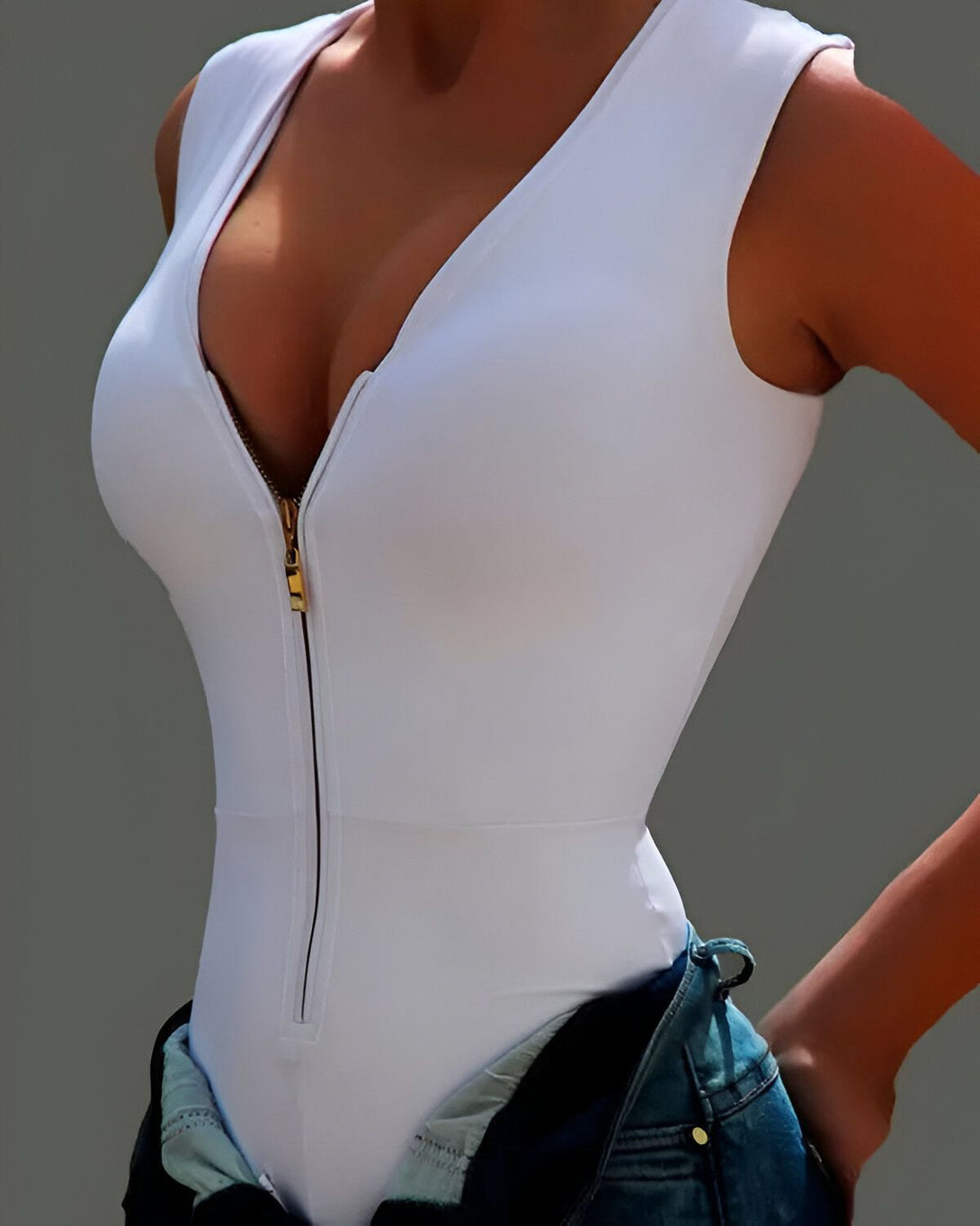 Une femme porte un body blanc moulant avec une fermeture éclair dorée sur le devant. Elle porte également un jean bleu.
