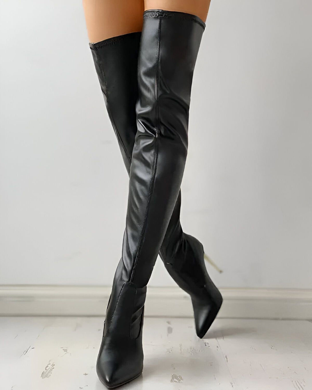 Une femme porte des bottes noires à talons hauts. Les bottes sont en cuir et ont une tige haute. La femme est debout et porte une robe noire.