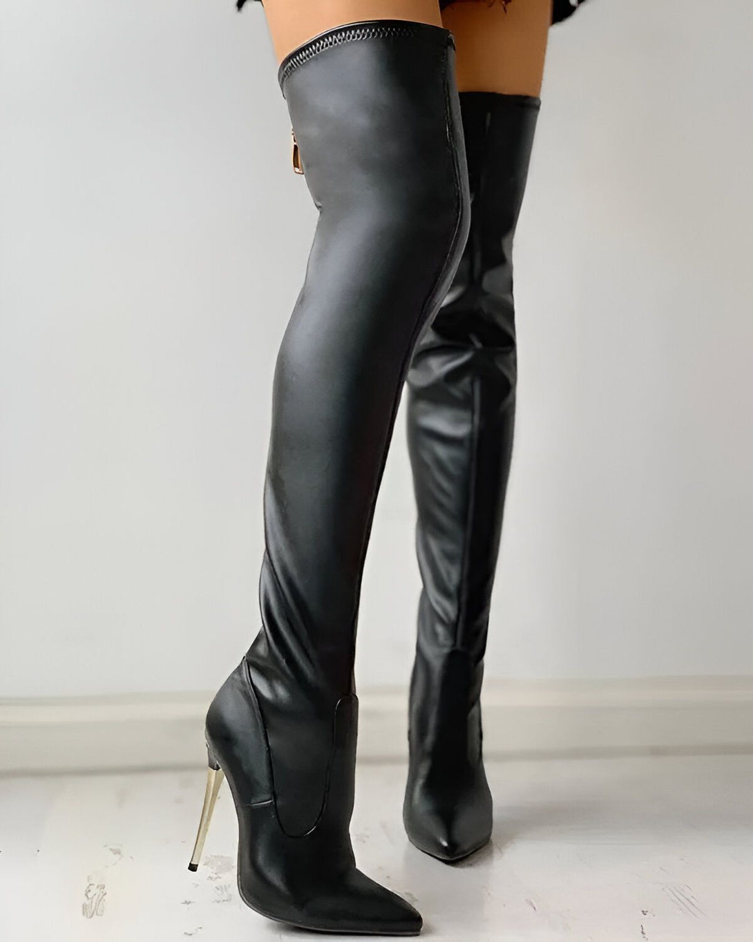 Une femme porte des bottes noires à talons hauts. Les bottes sont en cuir et ont une fermeture éclair sur le côté. La femme est debout et porte une jupe courte. On peut voir ses jambes et ses pieds. Les bottes sont très hautes et ont un talon fin. La femme est debout dans une pièce blanche. Il y a un mur blanc derrière elle.