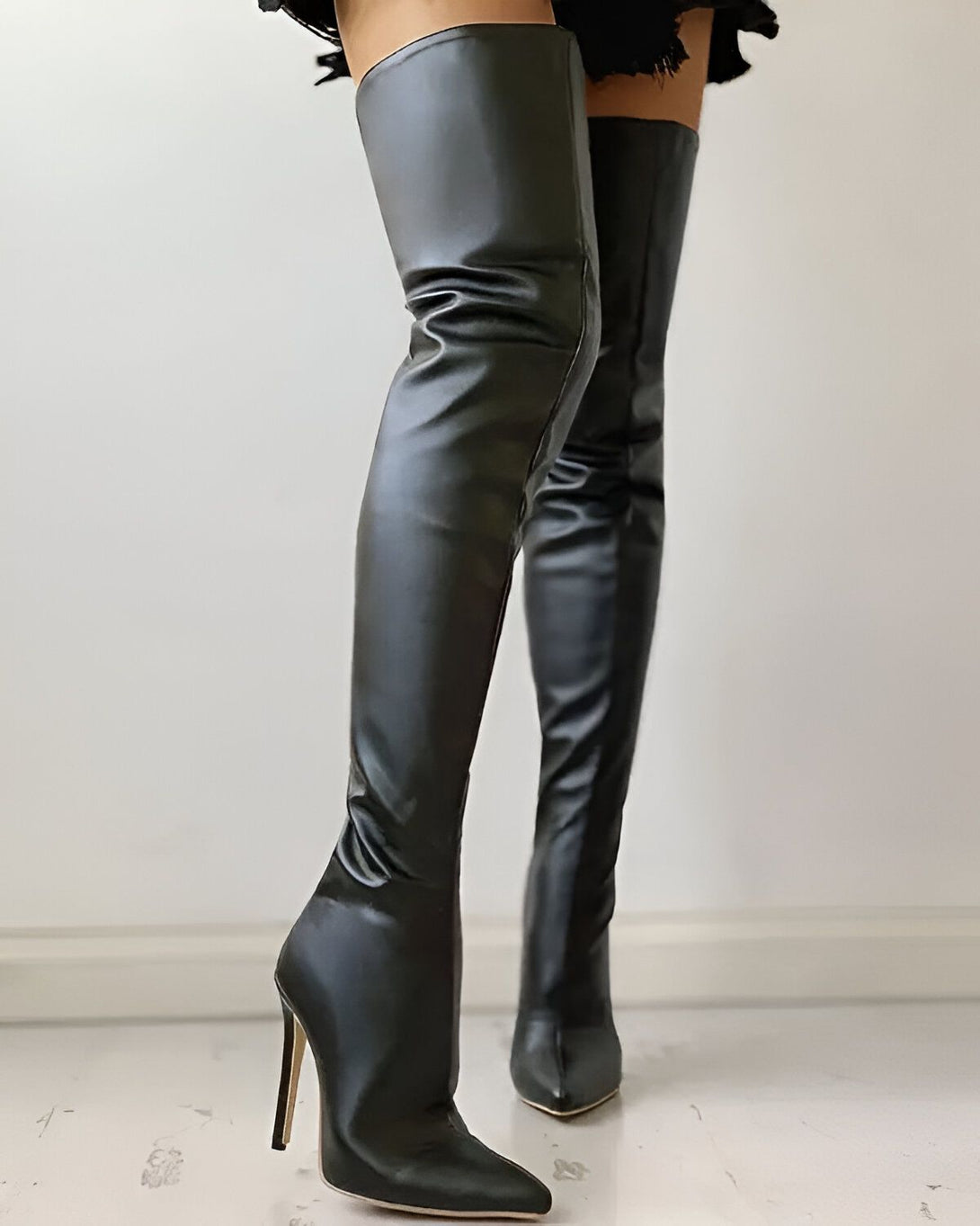 Une femme porte des bottes noires à talons hauts. Les bottes sont en similicuir et ont une tige haute. La femme est debout, les jambes légèrement écartées. Elle porte une jupe noire et un chemisier blanc. Ses cheveux sont longs et noirs. Elle est maquillée et porte des boucles d'oreilles.