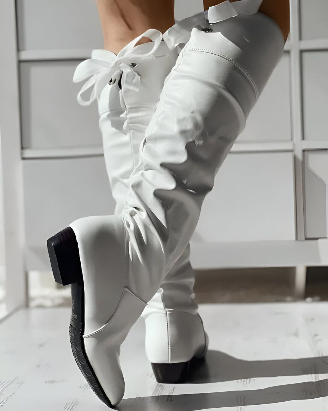 Une femme porte des bottes blanches à talons hauts. Les bottes sont en cuir et ont un bout pointu. La femme est debout sur un sol en bois.