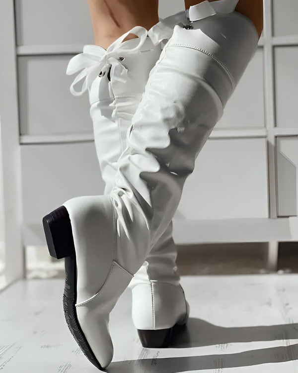 Une femme porte des bottes blanches à talons hauts. Les bottes sont en cuir et ont un bout pointu. La femme est debout sur un sol en bois.