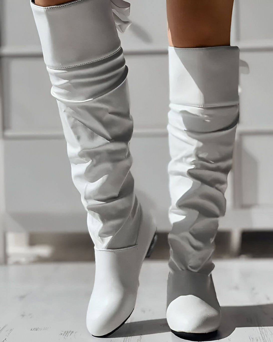 Une femme porte des bottes blanches à talons hauts. Les bottes sont en cuir et montent jusqu'au genou. La femme est debout sur un sol en carrelage beige. Elle porte une robe blanche et un manteau beige.