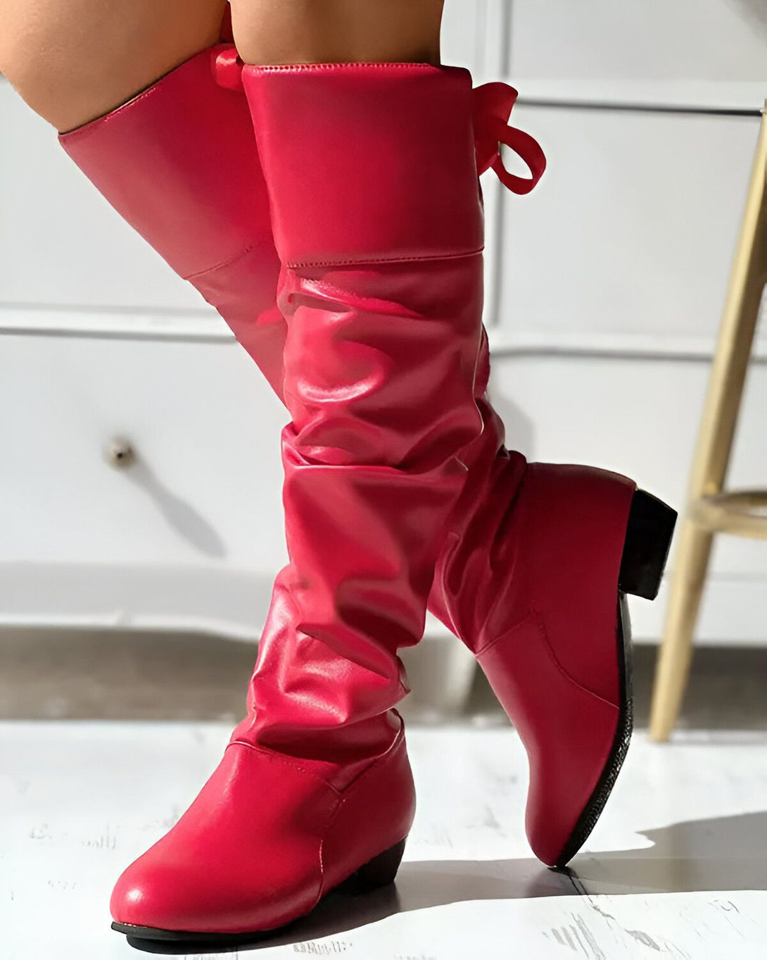 Une femme porte des bottes rouges à talons hauts. Les bottes sont en cuir et ont un bout pointu. La femme est debout sur un sol en bois.