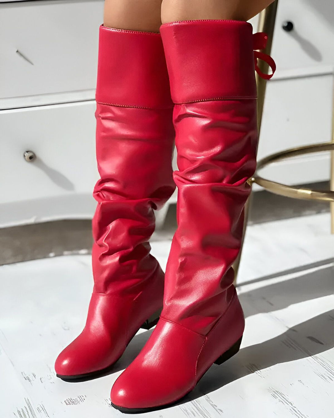 Une femme porte des bottes rouges à talons hauts. Les bottes sont en cuir et ont une tige froncée. La femme est debout sur un sol en bois.