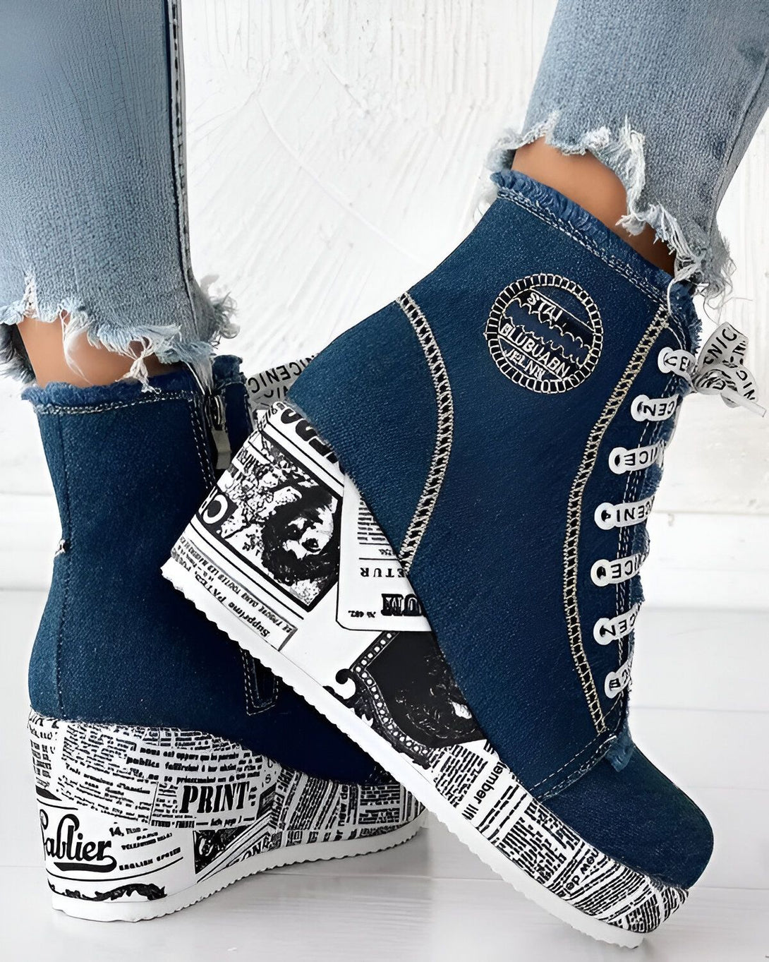 Une paire de bottines compensées en jean bleu avec un imprimé journal. Les bottines sont lacées sur le devant et ont une fermeture éclair sur le côté. Elles sont portées avec un jean bleu troué.