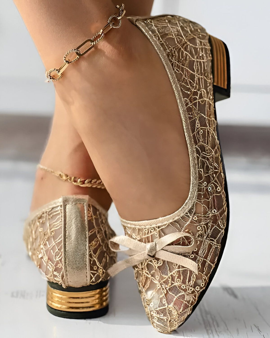 Une paire de chaussures plates dorées en dentelle avec un nœud sur le devant. Les chaussures sont portées par une femme qui a une chaîne dorée autour de la cheville.